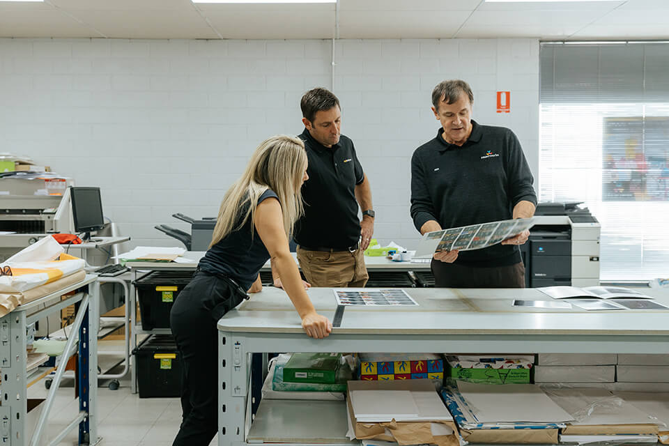 2 men and a woman looking at an A3 printed sheet of paper over a table